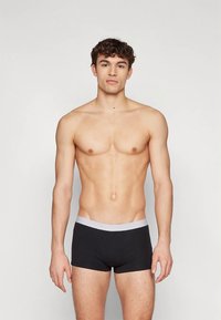 Melni bokseršorti ar pelēku jostasvietu, izgatavoti no elastīga auduma. Modelis stāv taisni pret vienkāršu fonu, demonstrējot piegriezumu un dizaina detaļas.