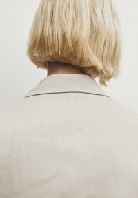 Veste beige clair avec une finition lisse et texturée et un col classique, vue de dos, complétée par de courts cheveux blonds.