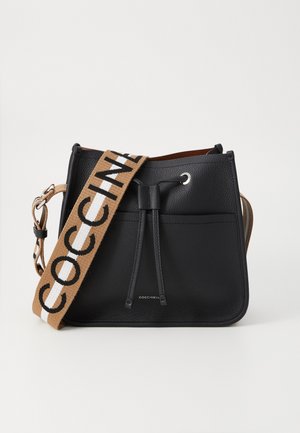 Black leather bucket bag with adjustable brown fabric strap featuring black lettering. Includes drawstring closure and front pocket.