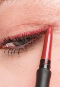 Crayon eye-liner rouge appliqué le long de la ligne des cils supérieurs, mettant en valeur une teinte rougeâtre scintillante sur un fond de peau neutre.