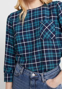 Chemise à carreaux avec une base marine, ornée de lignes turquoise et blanches. Comprend une poche poitrine avec un motif de carreaux en diagonale. Manches longues.