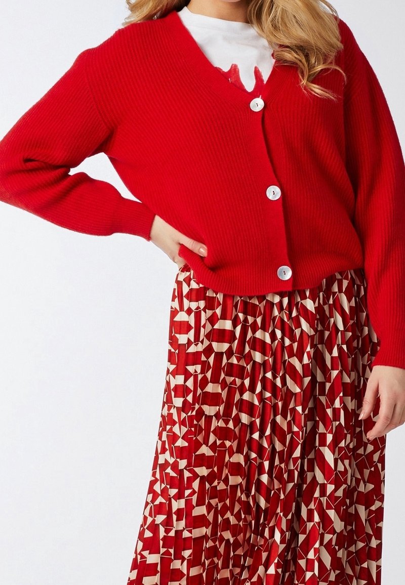 Czerwony kardigan prążkowany z białymi guzikami, zestawiony z plisowaną spódnicą z geometrycznym wzorem w odcieniach czerwieni i beżu.