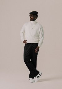 Maglione girocollo bianco, pantaloni sportivi neri, berretto nero, occhiali trasparenti, collane in oro a strati, sneaker bianche e nere. Sfondo neutro.