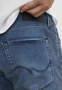 I jeans in denim presentano un lavaggio blu medio, una vestibilità dritta e una tasca posteriore con un dettaglio di cucitura curva, completata da una patta in pelle nera.