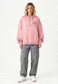 Felpa oversize rosa chiaro con grafica di una croce nera e testo, tasca frontale, abbinata a pantaloni cargo grigi e sneakers bianche.