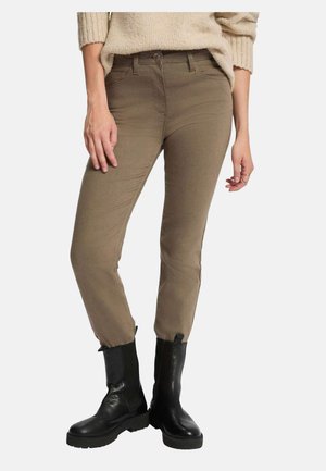 Pantalon marron slim porté avec un pull en tricot beige et des bottines en cuir noir sur une personne debout devant un fond blanc.