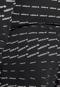 Svart tyg med ett upprepat vitt Adidas-logotypmönster. Designen inkluderar varierande orienteringar av logotypen och texten över materialet.