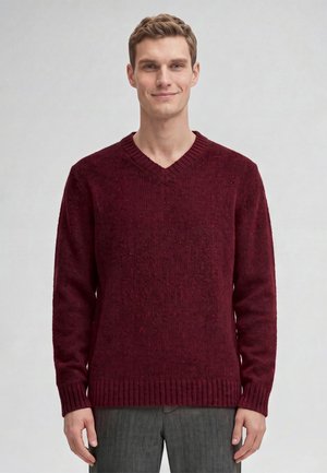 Giovane uomo che indossa un maglione a V color bordeaux e pantaloni grigi, in piedi e sorridente su uno sfondo chiaro e semplice.