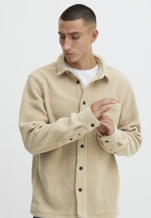 Beige fleece shirt jacket with button closure, notable texture, and black buttons. Features a collared neck and single chest pocket.
