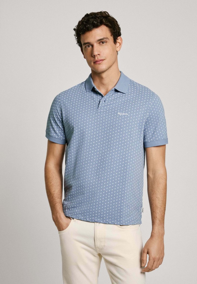 Mladý muž s kudrnatými vlasy, oblečený v modro vzorovaném polo tričku a krémových kalhotách, stojící s jednou rukou v kapse.