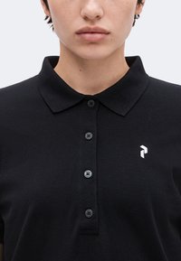 Gros plan d'une personne portant un polo noir avec un col, quatre boutons et un petit logo blanc sur le côté gauche de la poitrine.