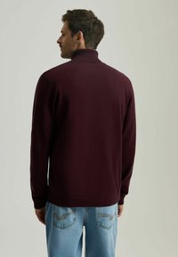 Bordowy sweter z wysokim kołnierzem, wykonany z miękkiego materiału, z długimi rękawami i dopasowanym krojem. Noszony z jasnoniebieskimi dżinsami.