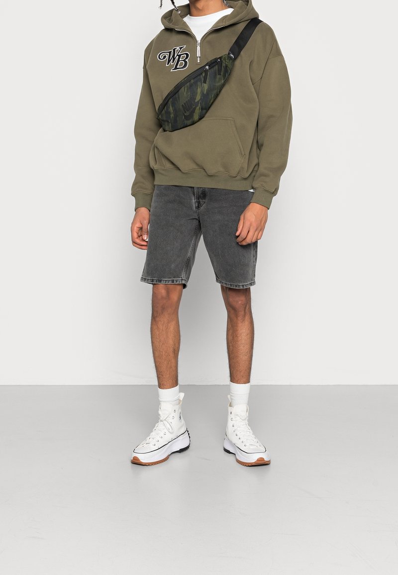 Sudadera con capucha color verde oliva y letras blancas, pantalones cortos de mezclilla gris y zapatillas blancas de caña alta. Bolsa de hombro de camuflaje negro y verde.