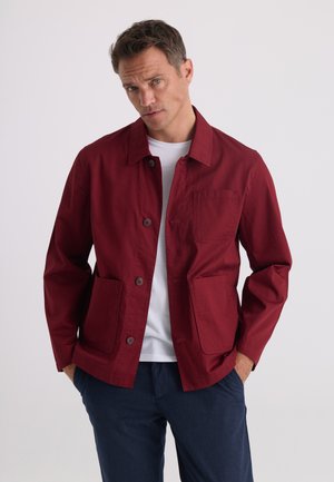 Homme portant une veste rouge boutonnée sur une chemise blanche et un pantalon bleu foncé, debout les mains dans les poches devant un fond uni.