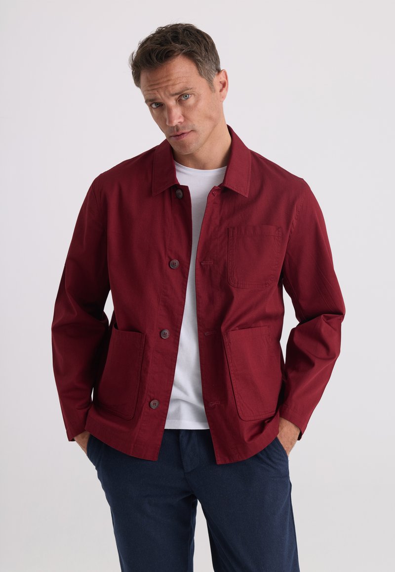 Homme portant une veste rouge boutonnée sur une chemise blanche et un pantalon bleu foncé, debout les mains dans les poches devant un fond uni.