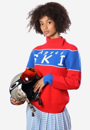 Roter und blauer Rollkragenpullover mit dem Text "Apres SKI", gerippte Textur. Das Modell hält einen silbernen Skihelm mit getöntem Visier.