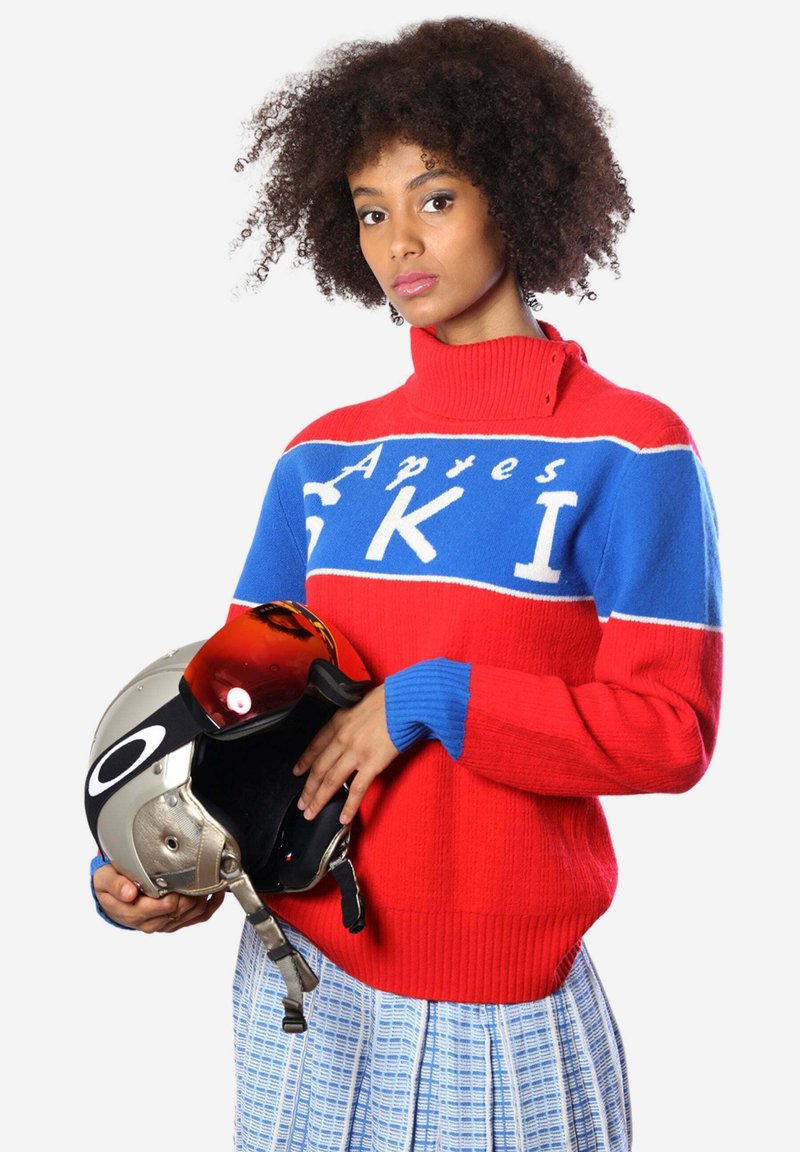 Maglione a collo alto rosso e blu con la scritta "Apres SKI", texture a coste. Il modello tiene in mano un casco da sci argento con visiera tinta.