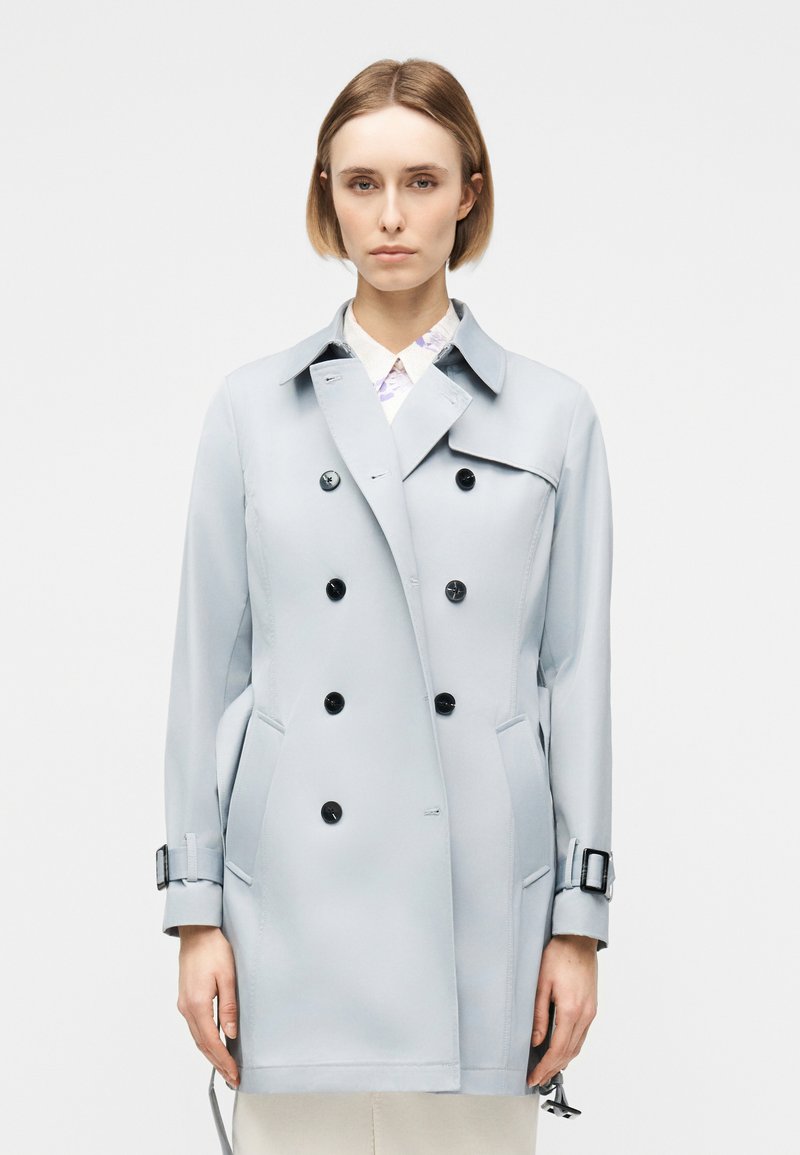 Γυναίκα με κοντά ξανθά μαλλιά που φοράει ένα ανοιχτό γκρι διπλόπετο παλτό τύπου καμπαρντίνα, στέκεται μπροστά σε ένα απλό λευκό φόντο.