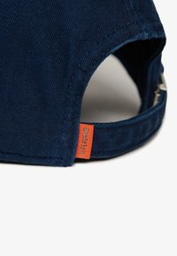 Gorra azul marino con tejido texturizado, que presenta una etiqueta naranja visible en el lateral, correa ajustable y visera curva.