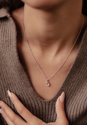Silver necklace with a delicate chain and a pear-shaped pink gemstone pendant, worn over a textured brown sweater.