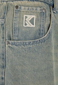 Vaaleansiniset farkut, joissa on näkyvä brodattu logo takataskussa, hopeinen nappi ja näkyviä ommeltuja yksityiskohtia.