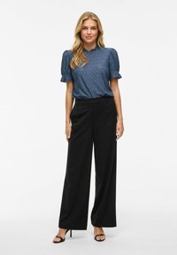 Blauwe gebloemde blouse met pofmouwen, gecombineerd met zwarte wijde broek en zwarte hoge sandalen. De blouse heeft een ronde hals.