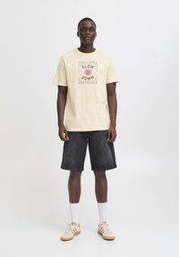 Beige bomulls t-skjorte med grafisk trykk i lilla og gult, paret med mørk denimshorts og hvite sokker, iført beige joggesko.