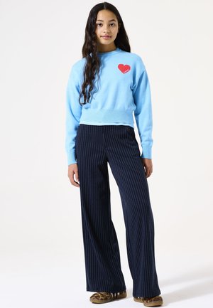 Un pull cropped bleu clair avec un détail en forme de cœur rouge sur la poitrine, associé à un pantalon large à rayures marines. Texture de tissu douce et coupe décontractée.