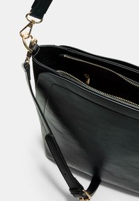 Sac à main en cuir noir avec une texture lisse, détails de fermeture éclair dorés et une bandoulière amovible. Il dispose de plusieurs compartiments intérieurs.