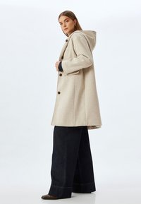 Abrigo de mezcla de lana beige con capucha, que cuenta con un cierre frontal de botones, grandes bolsillos laterales y un ajuste holgado, combinado con unos jeans oscuros de pierna ancha.