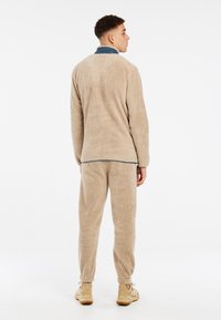 Giacca in pile beige soffice con colletto alto e pantaloni abbinati. Presenta polsini elastici e una vestibilità rilassata. Indossata con scarpe da ginnastica beige.