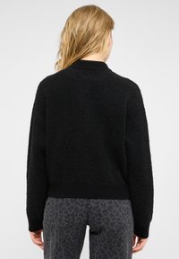 Pull noir en tissu pelucheux avec un col montant et un ourlet côtelé. Porté avec un jean gris à imprimé léopard, présentant une coupe décontractée et une longueur raccourcie.