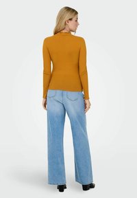 Mosterdgele geribbelde top met lange mouwen en een gekruide halslijn, gecombineerd met lichtblauwe wijde denim jeans en zwarte haklaarzen.