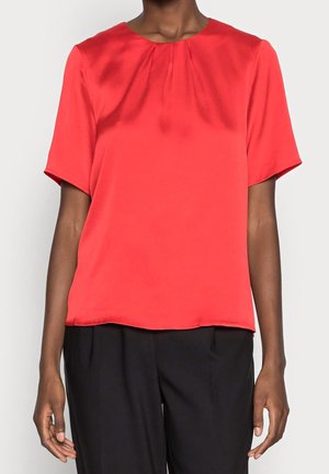Femme portant un chemisier rouge à manches courtes avec des plis près du col et un pantalon noir ample, debout devant un fond blanc.