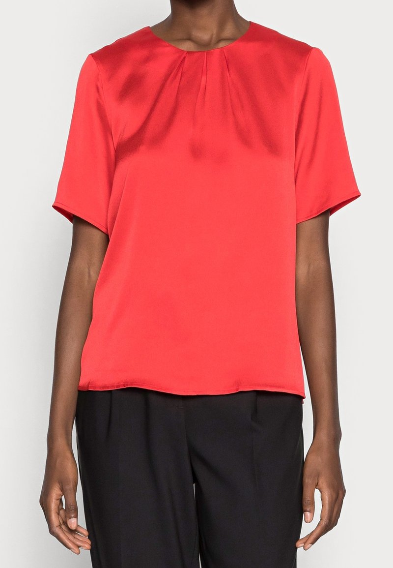 Femme portant un chemisier rouge à manches courtes avec des plis près du col et un pantalon noir ample, debout devant un fond blanc.