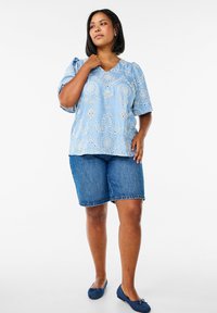 Blusa azzurra chiara con ricami floreali, maniche corte a palloncino e scollatura a V, abbinata a pantaloncini di jeans e scarpe ballerine blu.
