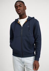 Marineblauwe rits-hoodie gemaakt van zacht materiaal, met een capuchon met trekkoorden, voorzakken en een contrasterende oranje voering.
