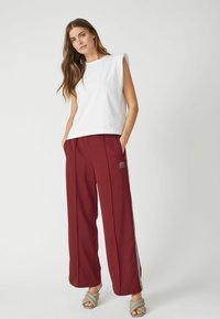 White sleeveless top with shoulder pads, paired with wide-leg burgundy trousers featuring side stripes and pockets. Soft, relaxed fit.