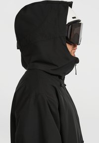 Zwarte hoodie buitenkleding met een gladde textuur, voorzien van een ritsluiting en een verstelbare trekkoordkap. Draagt een witte sk helmet en donkere goggles.