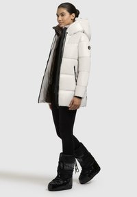 Giacca a vento bianca con cappuccio, cerniere nere e tasche laterali, abbinata a stivali da neve neri lucidi. Tessuto texture e design leggero.