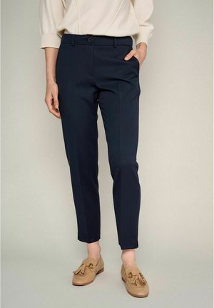 Pantalons navy ajustés avec une texture lisse, coupe slim, passants de ceinture et fermeture à un bouton. Associés à des chaussures à enfiler beige clair.