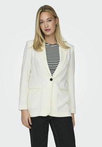 Hvid skræddersyet blazer med strukturerede skuldre, enkelt sort knap, to sidelommer og lange ærmer, båret over en sort og hvid stribet skjorte.