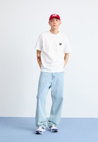 White short-sleeve t-shirt with a small black heart logo, paired with light blue baggy jeans and white sneakers with purple accents. Red cap.