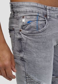 Jeans di denim grigio con una finitura testurizzata, caratterizzati da cuciture blu, un patch con logo e dettagli a coste unici sulla parte inferiore della gamba.