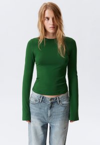 Grünes langärmliges, enganliegendes Oberteil aus dehnbarem Material, kombiniert mit hellblauen, distressed High-Waist-Jeans. Minimales Design und nahtlose Kanten.