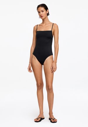 Femme debout portant un simple maillot de bain une pièce noir avec des bretelles fines et des tongs noires sur un fond blanc uni.