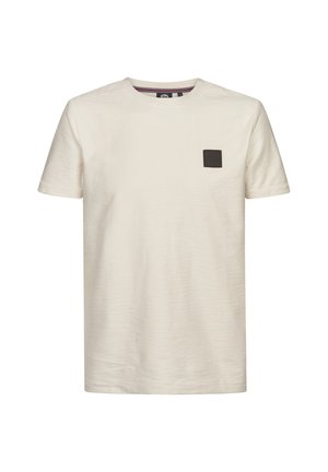 Camiseta color crema con cuello redondo, mangas cortas y tejido texturizado con un pequeño parche negro en el pecho izquierdo.