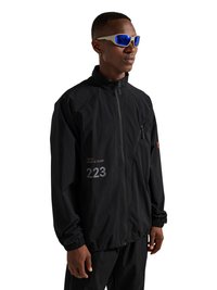 Man wearing black zip-up jacket with "223" and blue reflective sunglasses, standing against a white background.