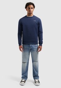 Donkerblauwe sweatshirt met geribde manchetten en een ronde halslijn, gecombineerd met lichtblauwe distressed jeans en zwart-witte sneakers.