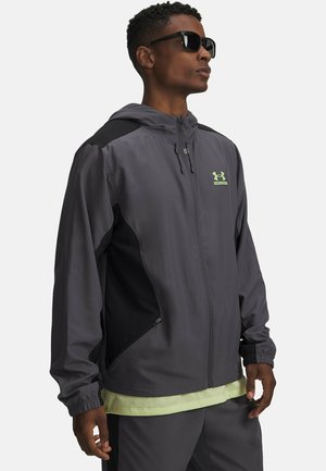 Graue Kapuzenjacke mit schwarzen Akzenten, ausgestattet mit einem Frontreißverschluss, elastischen Bündchen und einem reflektierenden Logo auf der Brust. Leichter Stoff.